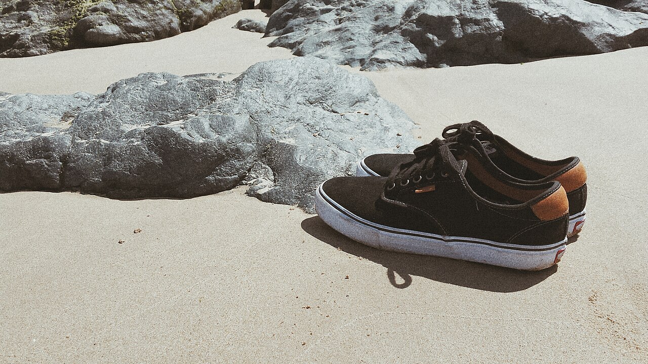 Black Vans shoes on sand beside beach rocks.